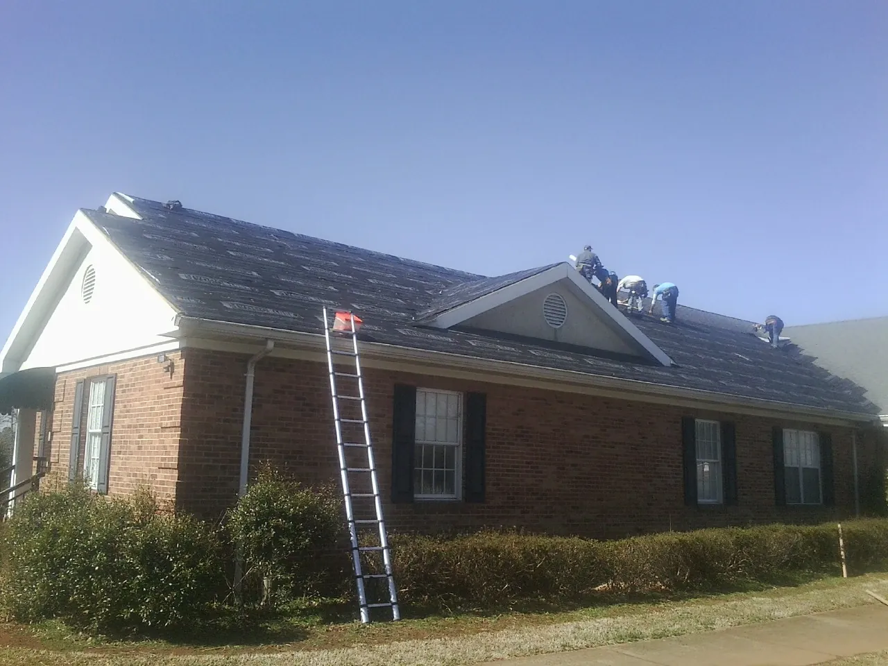 Crew tearing shingles off a brick ranch roof with a ladder set — Emergency Roof Repair work by our crew in Dellrose