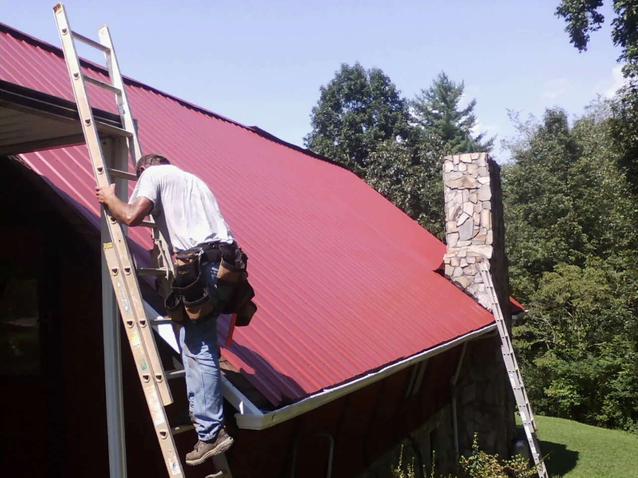 Roofer on a ladder beside a red metal roof and stone chimney — Soffit & Fascia Repair by Roofing Brothers in Dellrose